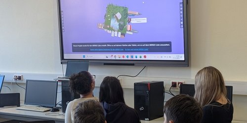 Fünf Kinder schauen auf einen Bildschirm, der über einem Tisch mit Computern hängt. 