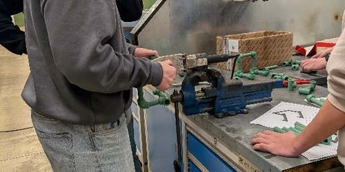 Ein Jugendlicher steht an einer Werkbank und hält ein grünes Rohr an eine Maschine.